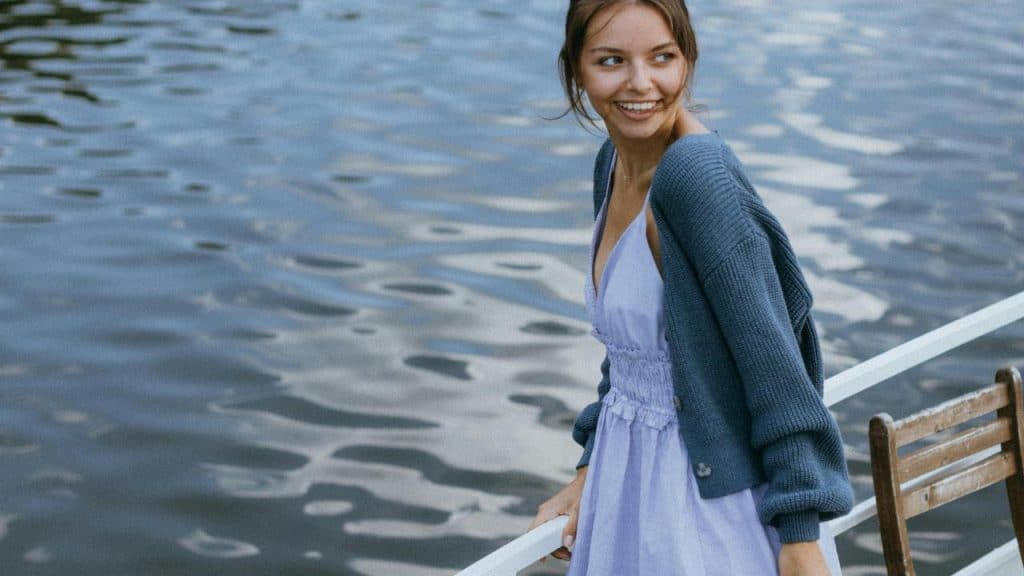 A smiling woman in a lavender dress and blue cardigan stands by the water.