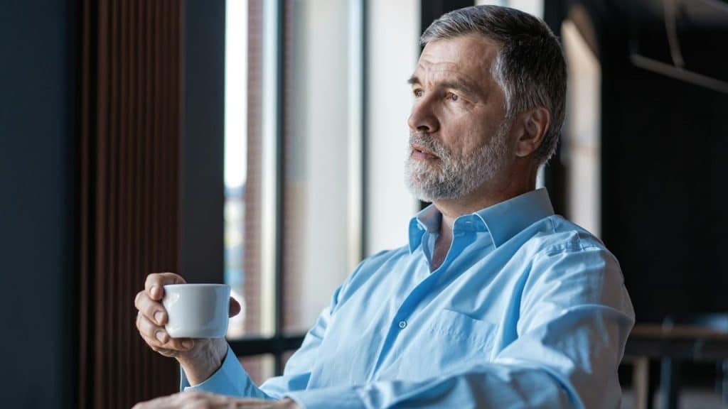 An older man with a grey beard holds a white cup while looking out a window.
