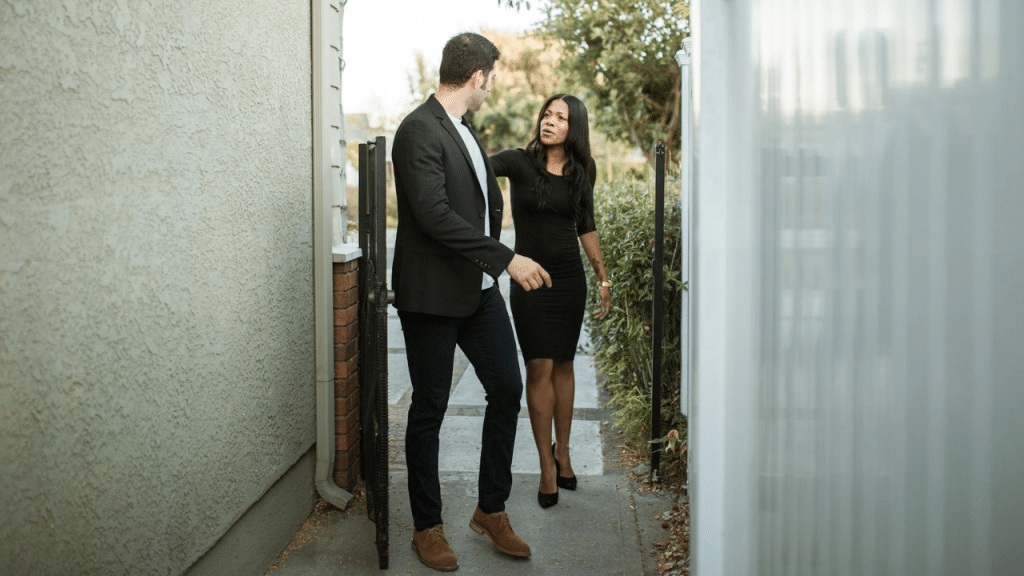 A woman in a black dress speaks to a man while standing near an outdoor gate.
