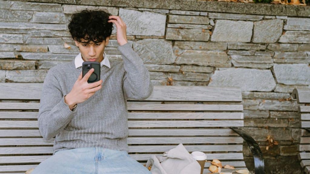A young man with curly hair sits on a park bench and looks at his smartphone.