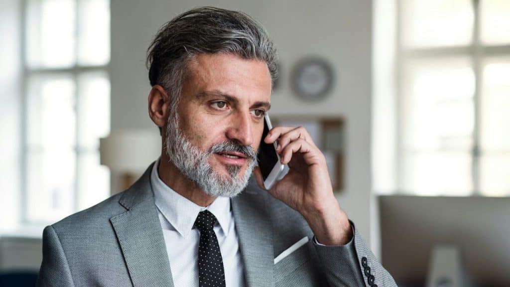A middle-aged man with gray hair and a beard wearing a suit talks on a smartphone.