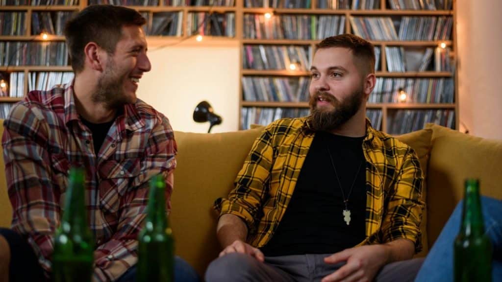 Two men in flannel shirts sit on a yellow couch and talk in a dimly lit room.