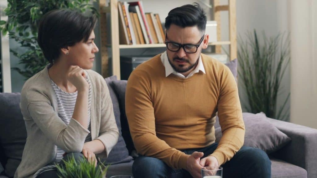 A woman in a cardigan and a man in a yellow sweater sit together on a couch.