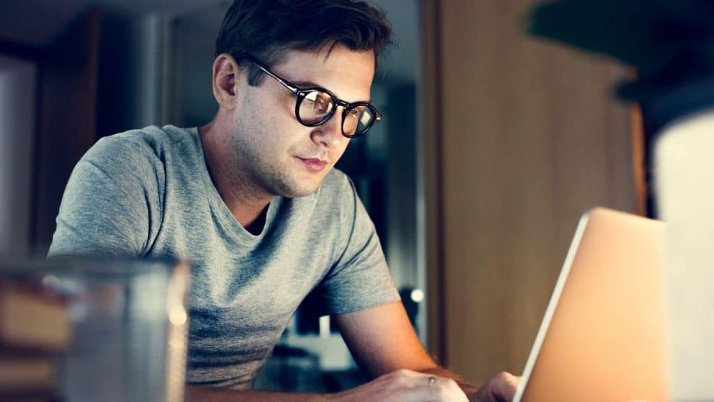 A man in a grey t-shirt and glasses looks intently at a glowing laptop screen.