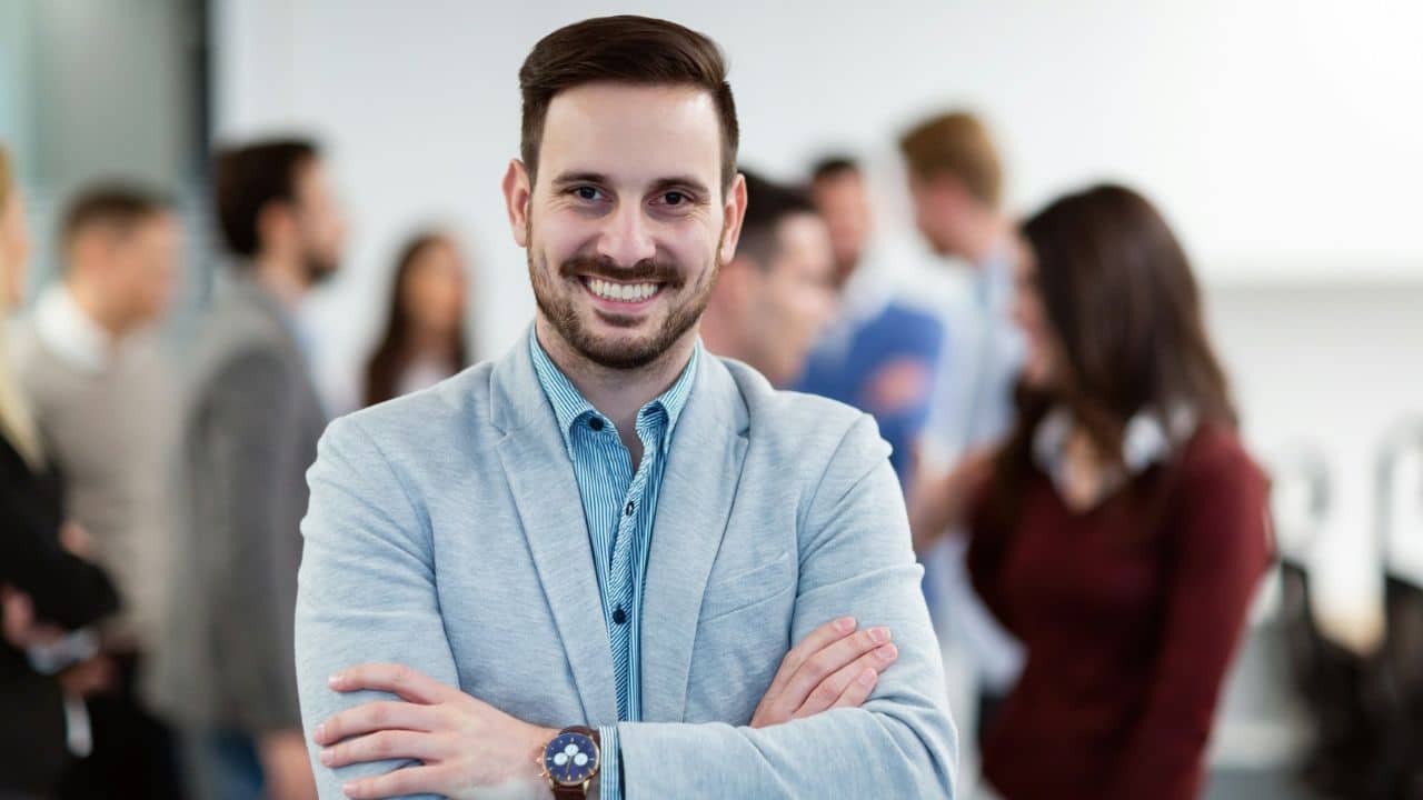 A smiling man in a blazer stands with arms crossed while people blur in background.
