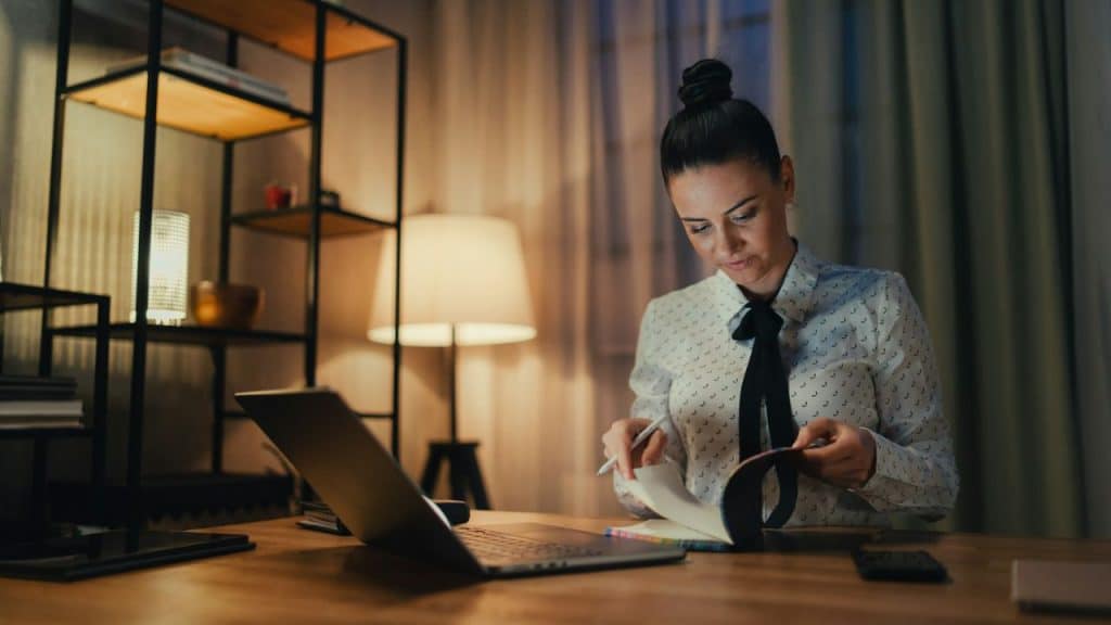 A woman with her hair in a bun sits at a desk at night, looking at a notebook next to a laptop.