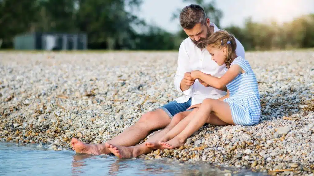 A man and his daughter sitting together on a pebble beach by the water.