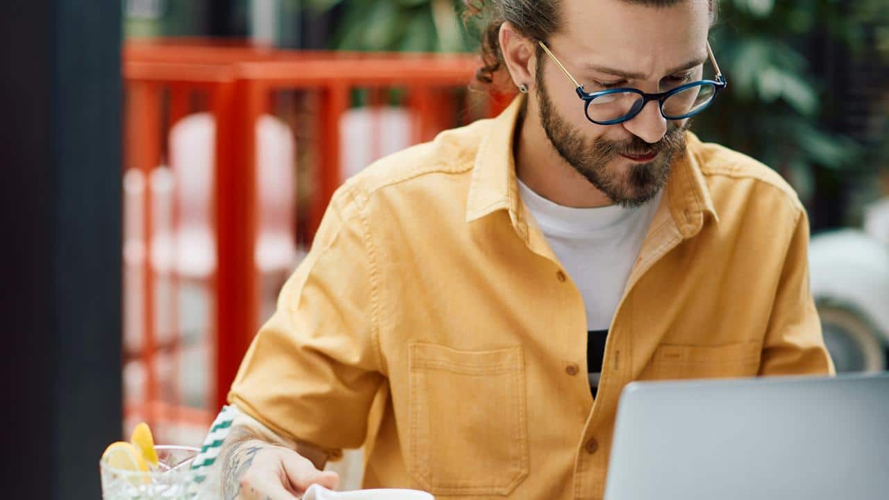 A man in a yellow shirt and glasses working on a laptop at a cafe.