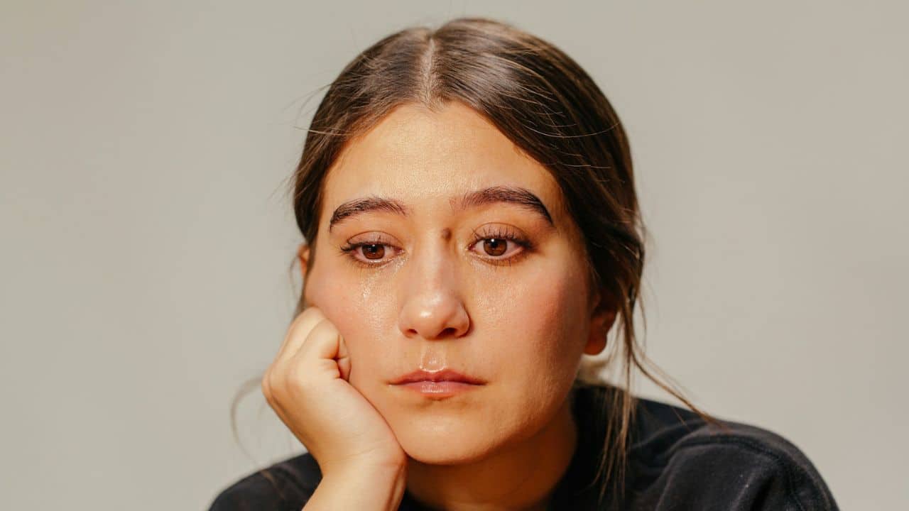 A woman with brown eyes rests her chin on her hand and looks down sadly.