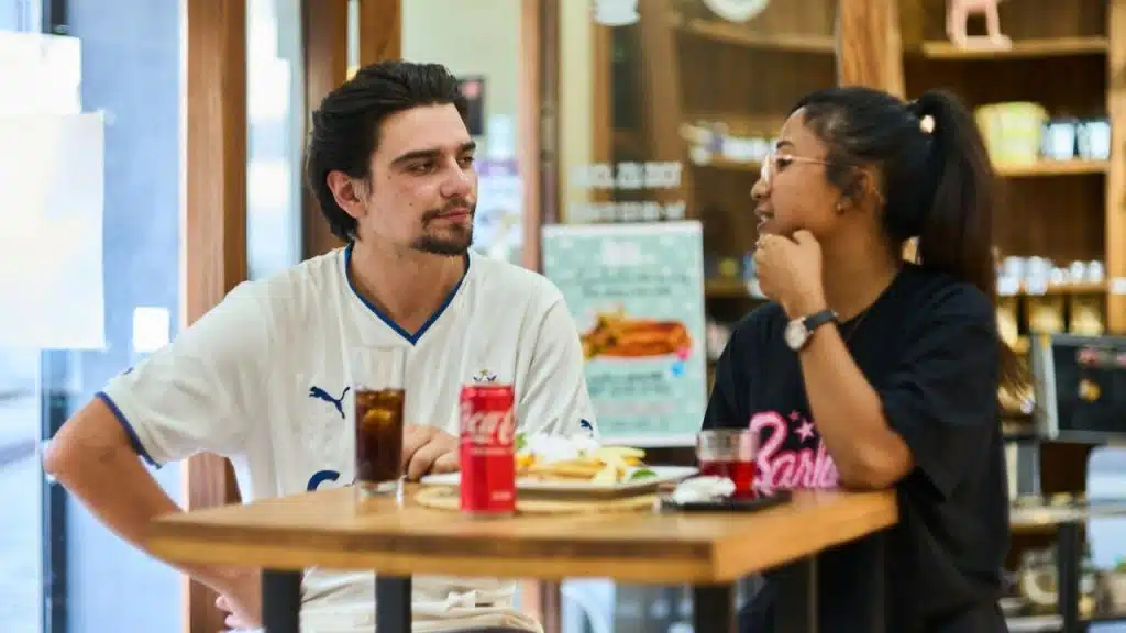 A man and woman sit at a cafe table with drinks, engaged in a conversation.