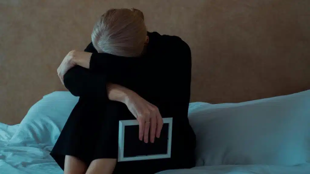 A woman in black sits on a bed with her head resting on her arms, holding a small picture frame in a somber setting.