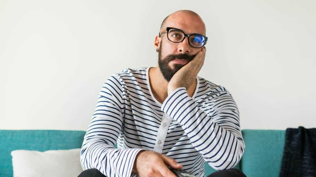 A bearded man wearing glasses is sitting on a couch and looking upset.