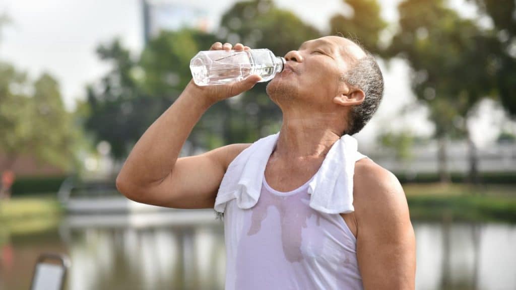 A sweaty man in a white tank top drinking water outdoors near a lake.