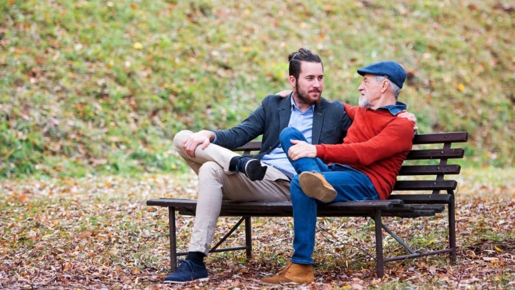 Two men sit on a park bench, one resting an arm on the other's shoulder.