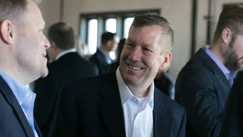 A middle-aged man in a dark suit and white shirt smiles while talking to another man in a crowded indoor