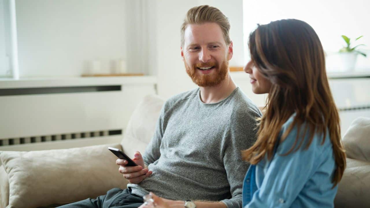 A man with red hair sits on a sofa holding a phone while talking to a woman.