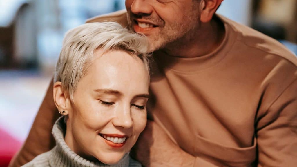 A close-up of a woman with short blonde hair smiling while a man leans in close to her, his chin resting near the top of her head.