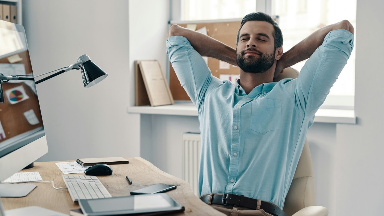 A man with a beard leans back in his office chair with hands behind his head.
