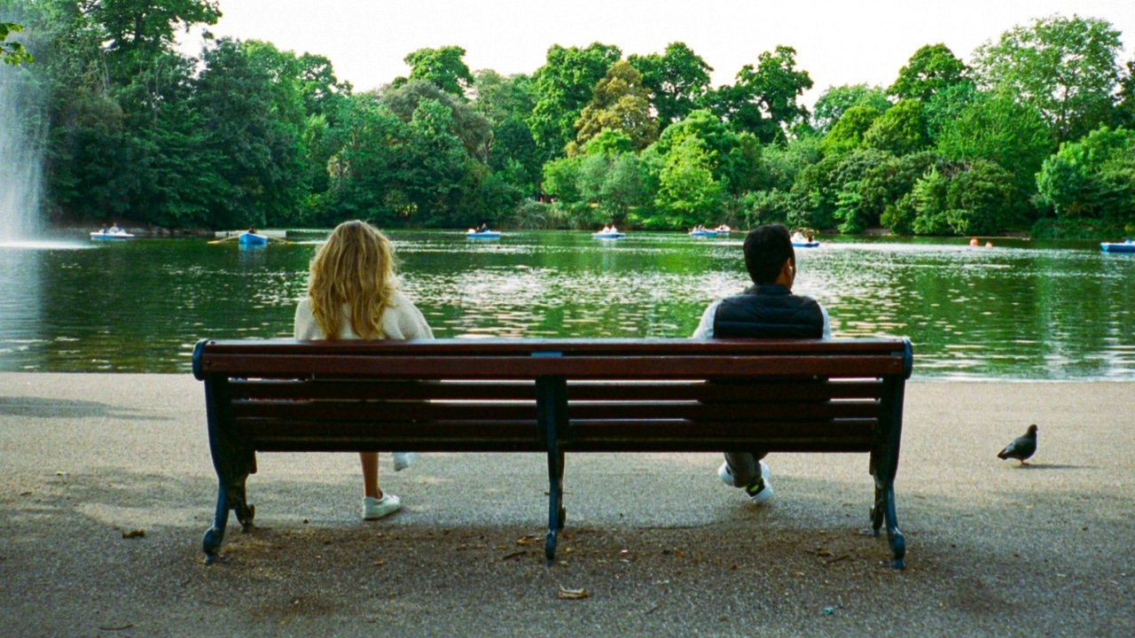 Two people sit on opposite ends of a park bench overlooking a pond with boats.