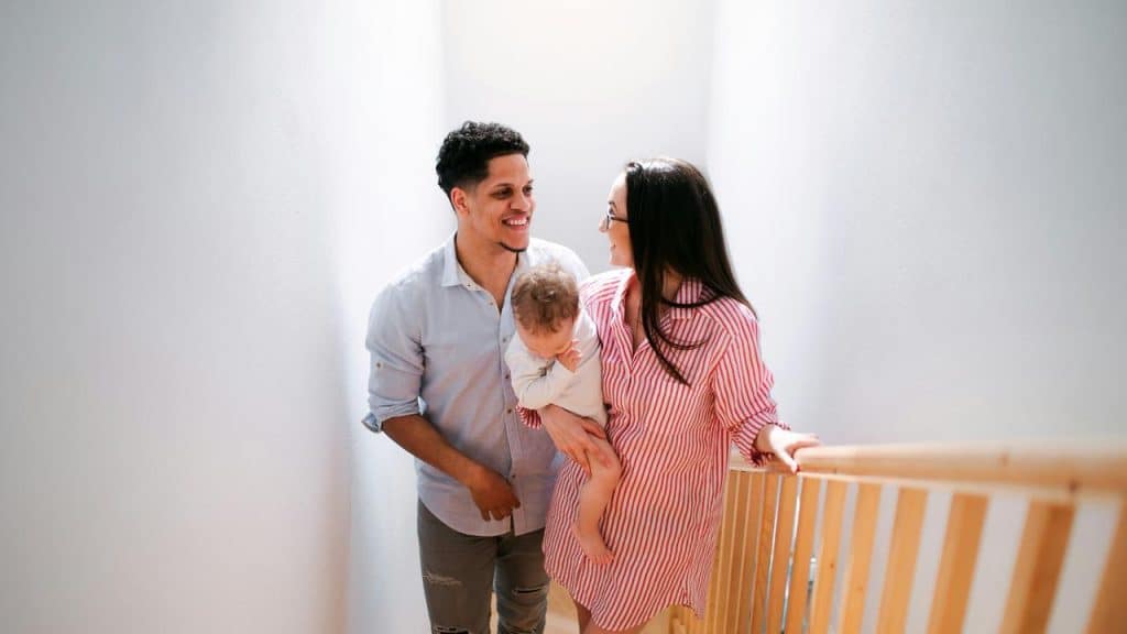 A smiling couple standing indoors while holding a baby near a staircase.