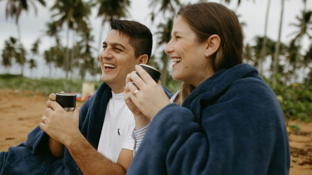 A man and woman laugh while holding mugs and sitting under a blanket on a beach.