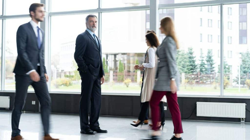 A man in a suit stands still in a bright lobby as people walk past.
