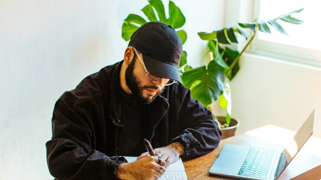A man wearing a black cap and glasses writes in a notebook beside a laptop.