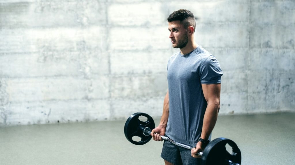 A man in a gray athletic shirt lifts a weighted barbell in a gym setting.