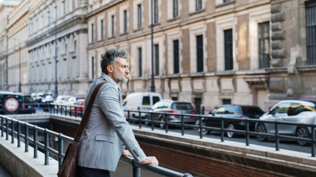 A man in a grey blazer stands by a railing overlooking a city street.