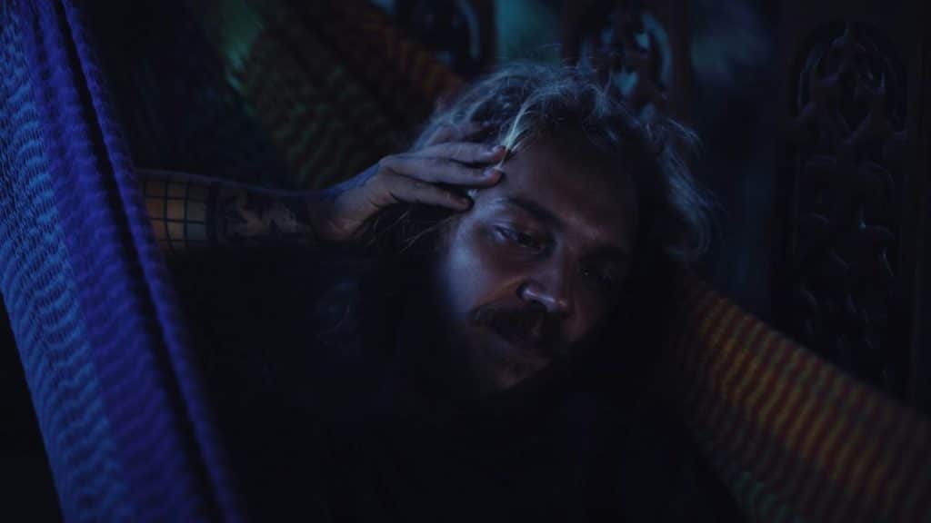 A man with a mustache rests in a colorful hammock, holding one hand to his forehead.