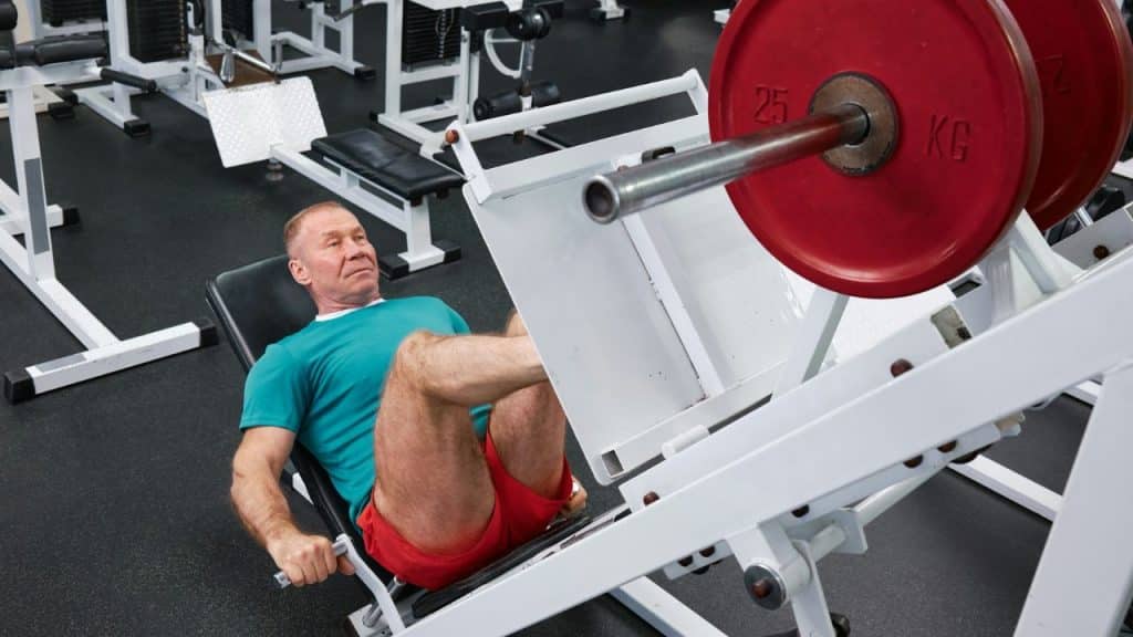 A man in athletic wear uses a leg press machine with heavy red weights.