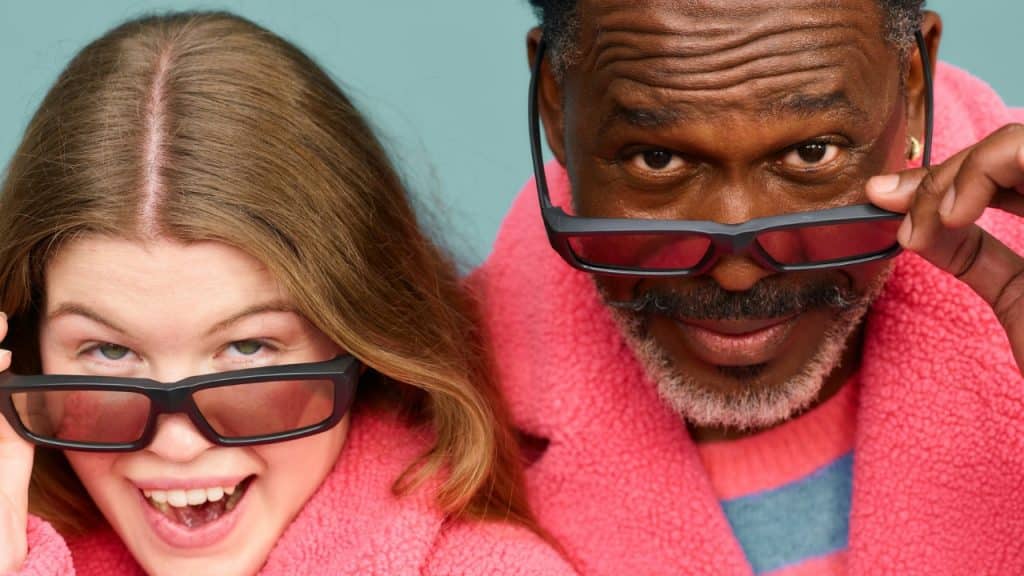 A woman and a man wearing pink fuzzy coats peer over their dark-framed glasses.