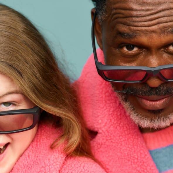 A woman and a man wearing pink fuzzy coats peer over their dark-framed glasses.