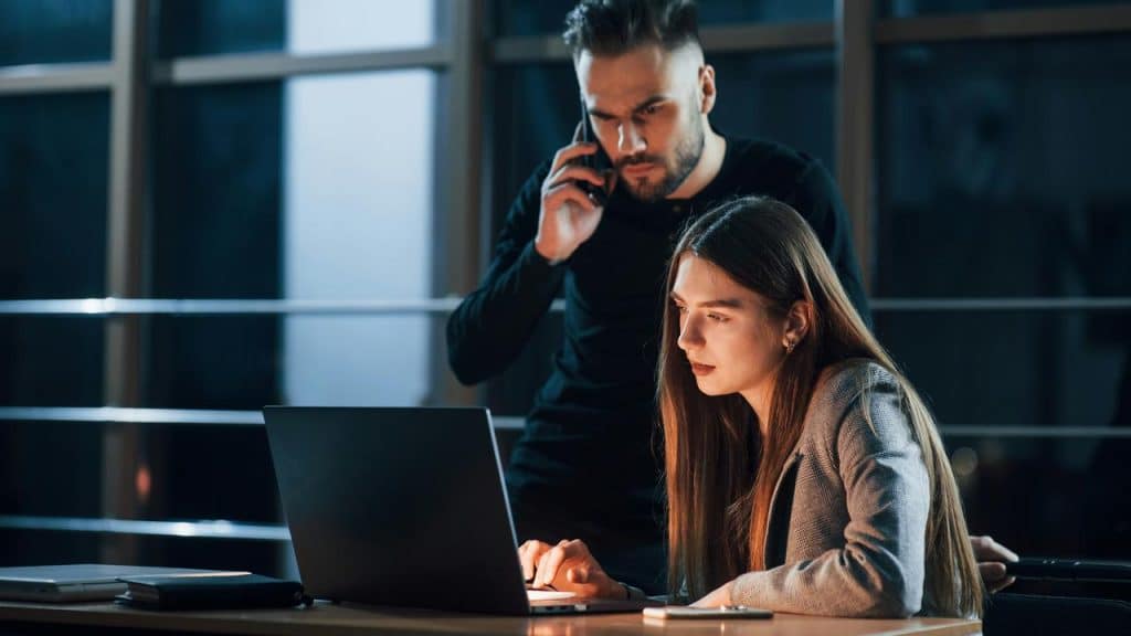 A man on the phone stands behind a woman focused on a laptop.