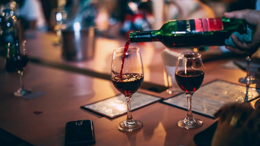 A bottle pouring red wine into glasses on a bar counter.