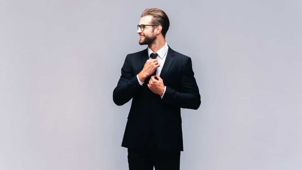 A smiling man in a black suit and glasses adjusts his necktie against a grey background.