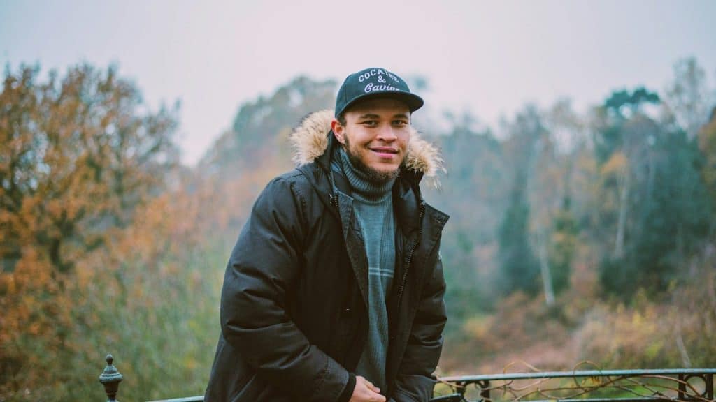A smiling man wearing a parka in front of a handrail and trees.