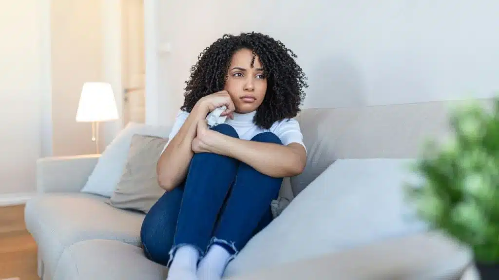 A woman with curly hair sits on a couch, hugging her knees and looking away.