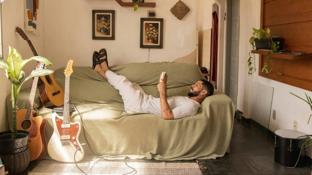 A man looking at his phone while lying on a couch in his living room while two guitars stand by the couch’s end.