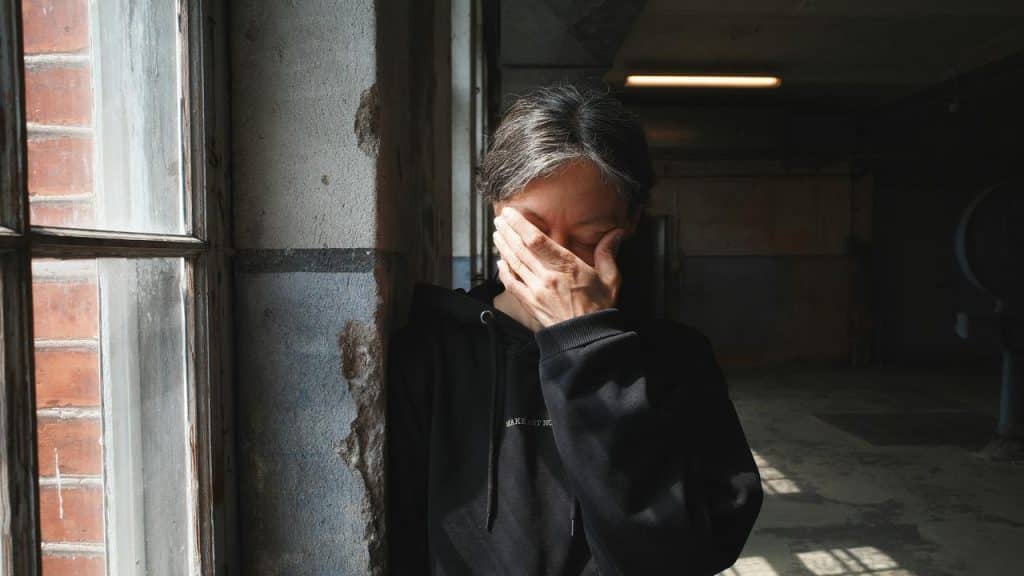 A person standing by a window in a dim hallway, covering their face with one hand.