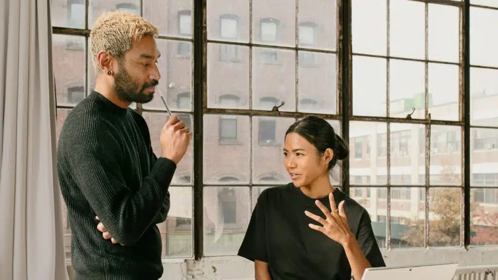 A man with blonde hair and a woman in black shirts converse in front of a window.