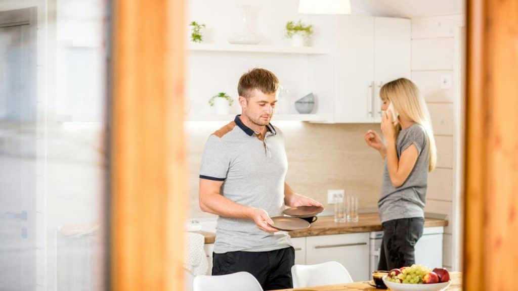 A man holds two plates in a kitchen while a woman talks on the phone in the background.