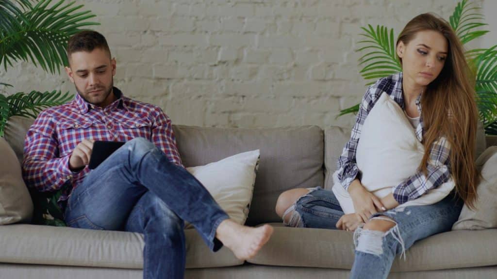 A woman in a plaid shirt and a man in a checkered shirt sit apart on a sofa.