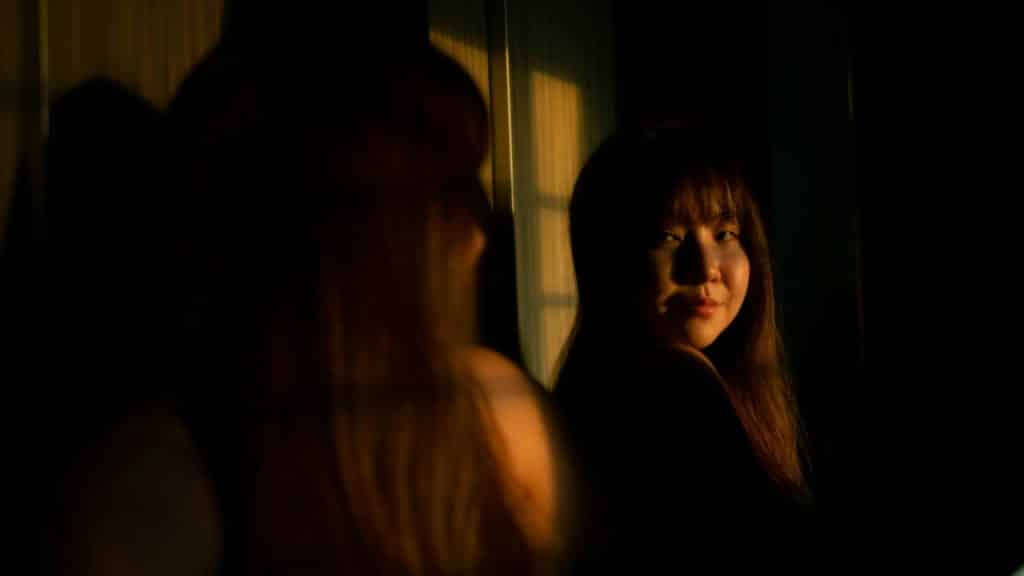 A woman looks into a mirror at her own reflection in a dimly lit room.