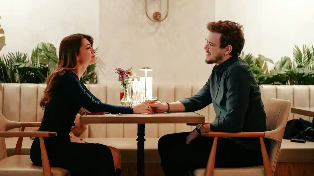 A man and woman sit at a table, smiling at each other while holding hands.