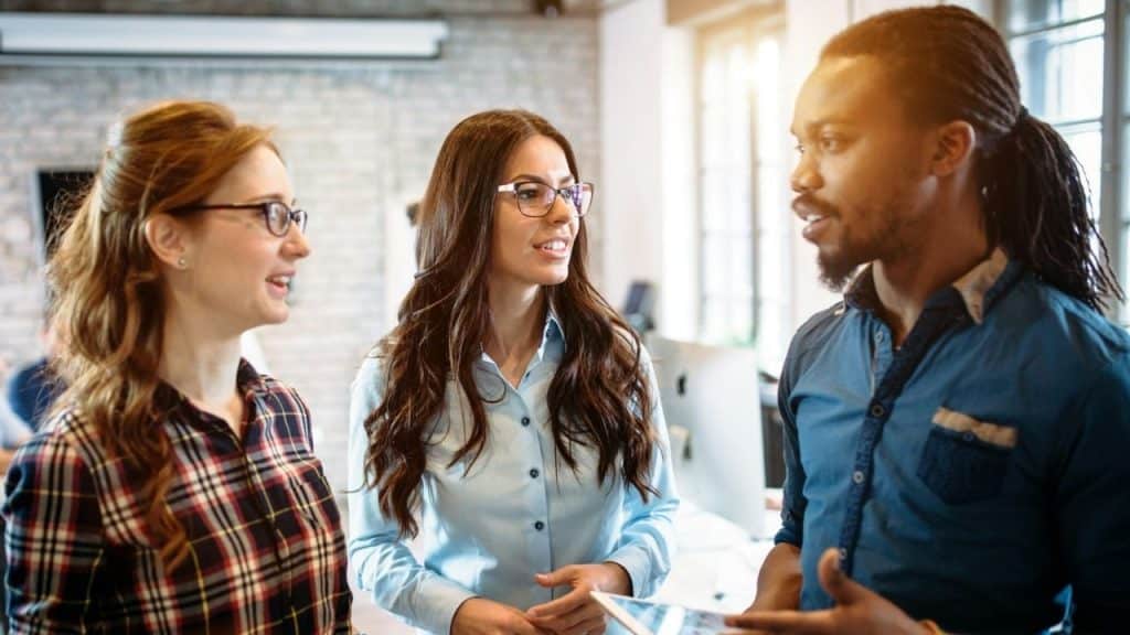 Two women stand in an office setting and listen to a man who is speaking.