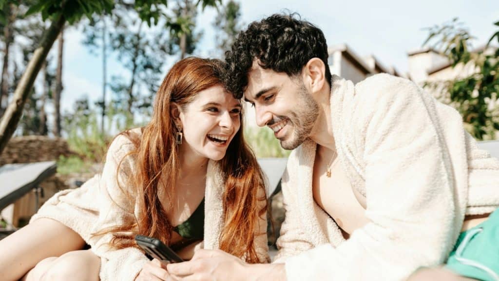 A smiling man and woman in robes sit outside while looking at a mobile phone.