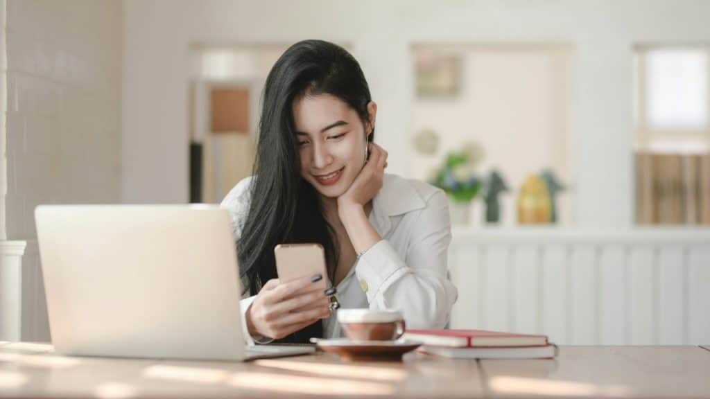 A smiling woman looks at her phone while sitting at a table with a laptop.