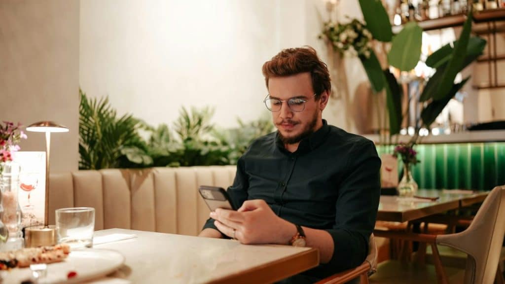 A man in a dark green shirt and glasses looks down at his phone in a restaurant.