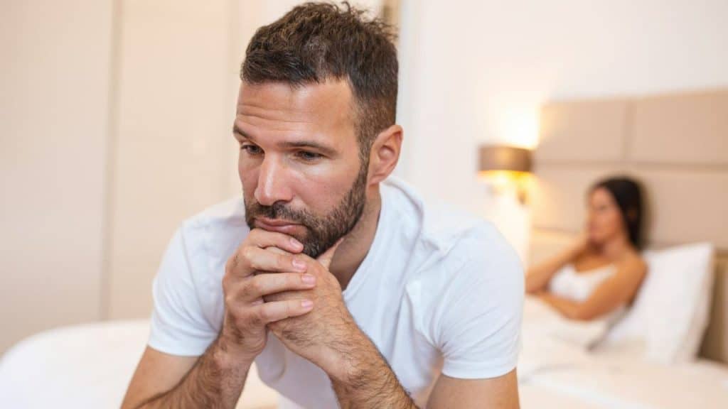 A man sits on a bed with his chin resting on his clasped hands.